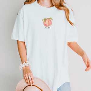 May include: A white oversized t-shirt with a peach graphic and the word "peachy" printed on the front.