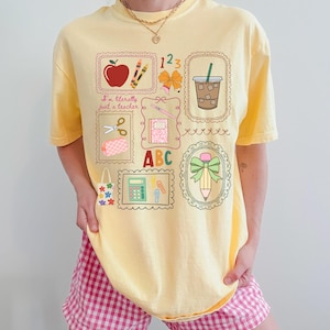 May include: Yellow t-shirt with a colorful teacher-themed graphic. The graphic includes an apple, pencils, scissors, a notebook, a calculator, a pencil, and a coffee cup. The text "I'm literally just a teacher" is also included.