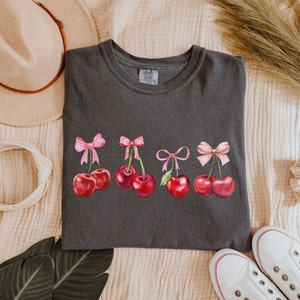 Puede incluir: Una camiseta gris oscuro con un gr&aacute;fico de cinco cerezas rojas con lazos rosas. Las cerezas est&aacute;n dispuestas en una fila a trav&eacute;s del centro de la camiseta.