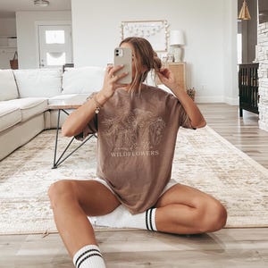 May include: A brown oversized t-shirt with a white graphic of wildflowers and the text "wild like wildflowers". The model is wearing the shirt with white shorts and white socks.