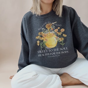 Puede incluir: Una sudadera gris oscuro con un gr&aacute;fico de un tarro de miel con una abeja y flores. El texto en la sudadera dice "Kind words are like honey, Sweet to the soul, Healthy for the body, Proverbs 16:24".