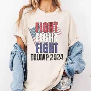 May include: A white t-shirt with a red, white, and blue American flag graphic and the text "FIGHT FIGHT FIGHT TRUMP 2024".