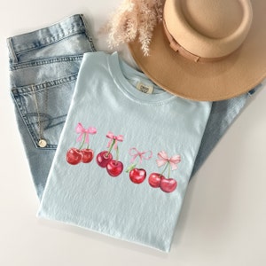 Puede incluir: Una camiseta azul claro con un gr&aacute;fico de cinco cerezas rojas con lazos rosas en los tallos. Las cerezas est&aacute;n dispuestas en una fila a trav&eacute;s de la parte delantera de la camiseta.