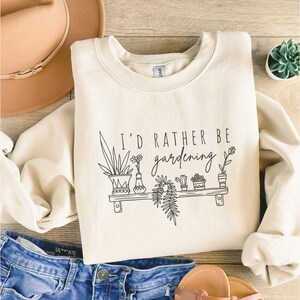Peut inclure: Un sweat-shirt cr&egrave;me avec le texte "I'd rather be gardening" et un dessin au trait noir de plantes en pot sur une &eacute;tag&egrave;re.