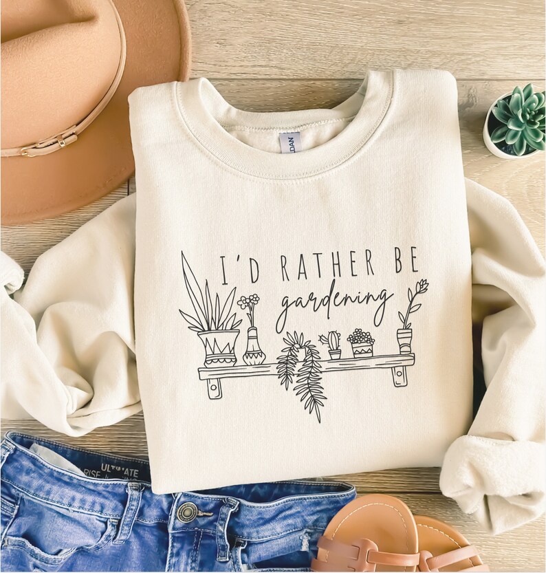 Peut inclure: Un sweat-shirt cr&egrave;me avec le texte "I'd rather be gardening" et un dessin au trait noir de plantes en pot sur une &eacute;tag&egrave;re.