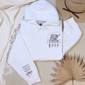 Pode incluir: Camisola de capuz branca com um gr&aacute;fico preto e branco de um peixe com iscas de pesca penduradas. O texto "CUSTOM" est&aacute; impresso no gr&aacute;fico. A manga tem um gr&aacute;fico preto e branco de um peixe com o texto "HAILEY CARLEY BENNY".