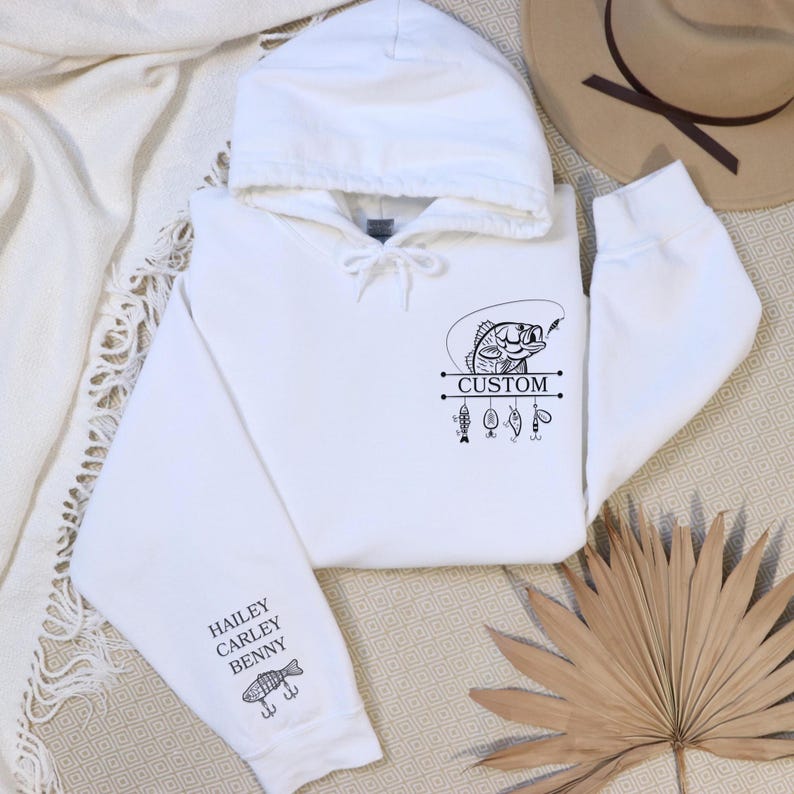 Pode incluir: Camisola de capuz branca com um gr&aacute;fico preto e branco de um peixe com iscas de pesca penduradas. O texto "CUSTOM" est&aacute; impresso no gr&aacute;fico. A manga tem um gr&aacute;fico preto e branco de um peixe com o texto "HAILEY CARLEY BENNY".