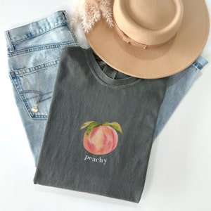 May include: A dark gray t-shirt with a peach illustration and the word "peachy" printed in white.