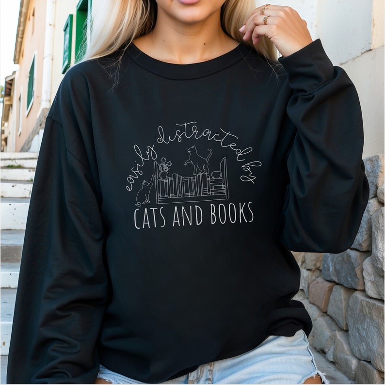 May include: Black sweatshirt with a white graphic design that says "easily distracted by cats and books". The graphic features a bookshelf with cats and plants.