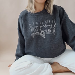 Peut inclure: Un sweat-shirt gris fonc&eacute; avec le texte "I'd rather be gardening" en lettres blanches. Le texte est accompagn&eacute; d'un dessin au trait de plantes en pot sur une &eacute;tag&egrave;re.