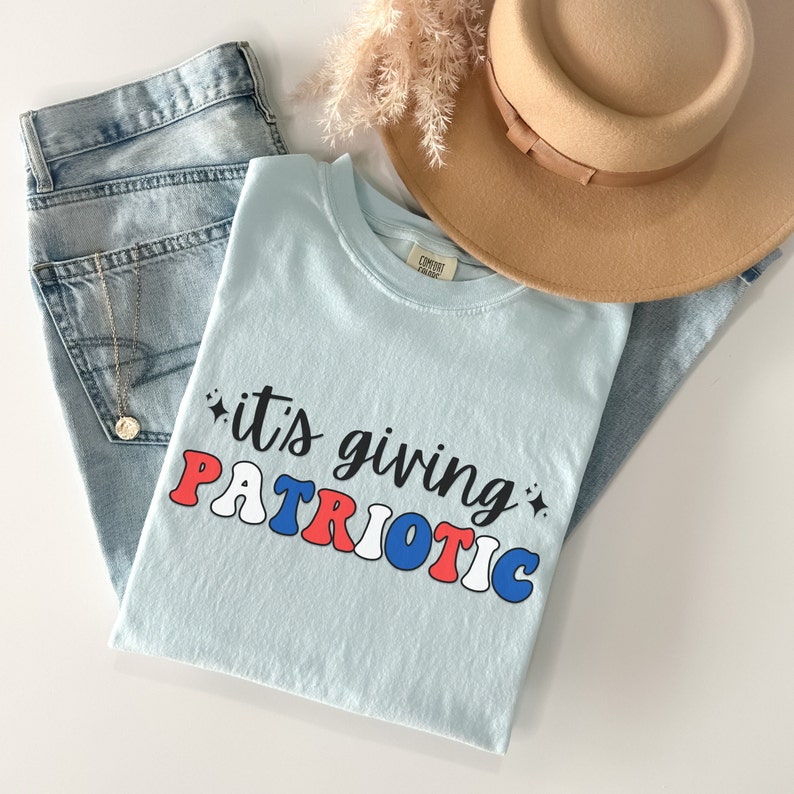 Pu&ograve; includere: Una maglietta azzurra con la scritta "it's giving PATRIOTIC" in rosso, bianco e blu. La maglietta &egrave; appoggiata su un paio di jeans blu chiaro, con un cappello marrone e una collana d'oro. Nell'immagine ci sono anche fiori secchi.