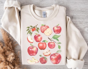 May include: A beige sweatshirt with a watercolor design of red apples and leaves. The apples are in various stages of ripeness, some whole and some sliced.