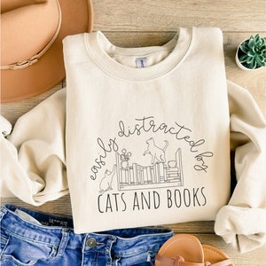 May include: A cream-colored sweatshirt with the text "easily distracted by cats and books" in black. The text is surrounded by a circle and a drawing of two cats near a bookshelf.