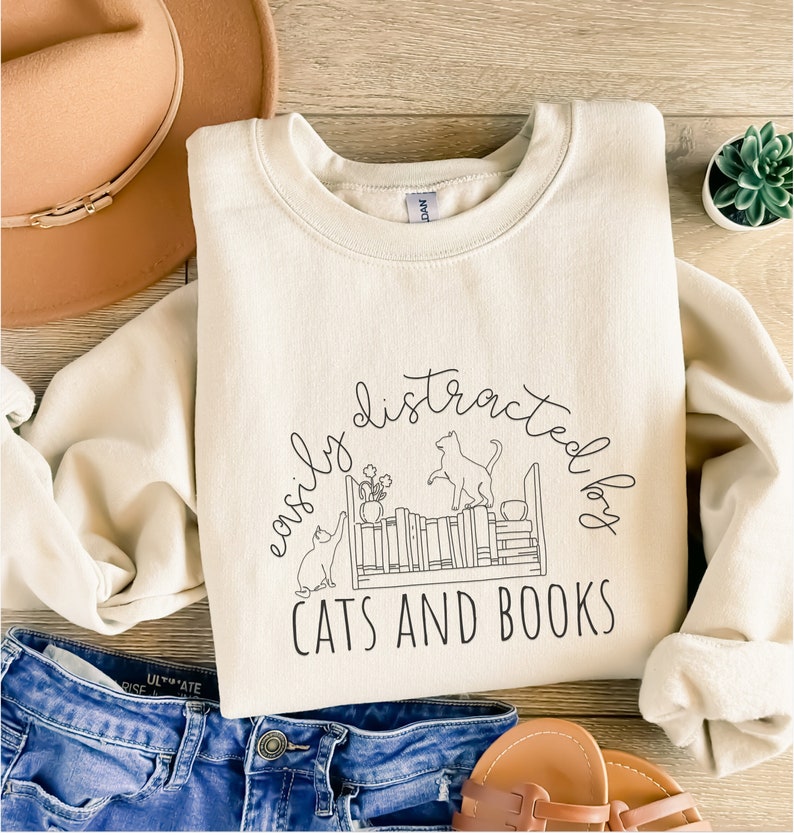 May include: A cream-colored sweatshirt with the text "easily distracted by cats and books" in black. The text is surrounded by a circle and a drawing of two cats near a bookshelf.
