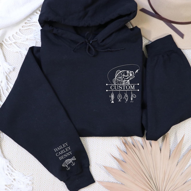 Pode incluir: Camisola de capuz preta com um gr&aacute;fico branco de um peixe com uma linha de pesca e a palavra "CUSTOM" numa faixa. A manga tem os nomes "HAILEY, CARLEY, BENNY" e um pequeno gr&aacute;fico de peixe.