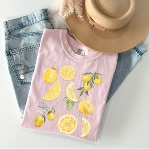 Pu&ograve; includere: Una maglietta rosa con un design ad acquerello di limoni e foglie. La maglietta ha un collo rotondo e maniche corte. Il testo "CONFETTI CLOUDS" &egrave; stampato sulla maglietta.