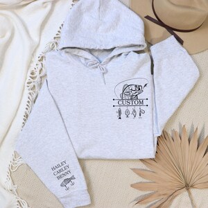 Pode incluir: Camisola de moletom cinza com capuz com um gr&aacute;fico preto de um peixe em uma linha de pesca com a palavra "CUSTOM" e outros iscas de pesca. A manga tem os nomes "HAILEY, CARLEY, BENNY" e um gr&aacute;fico de um peixe.