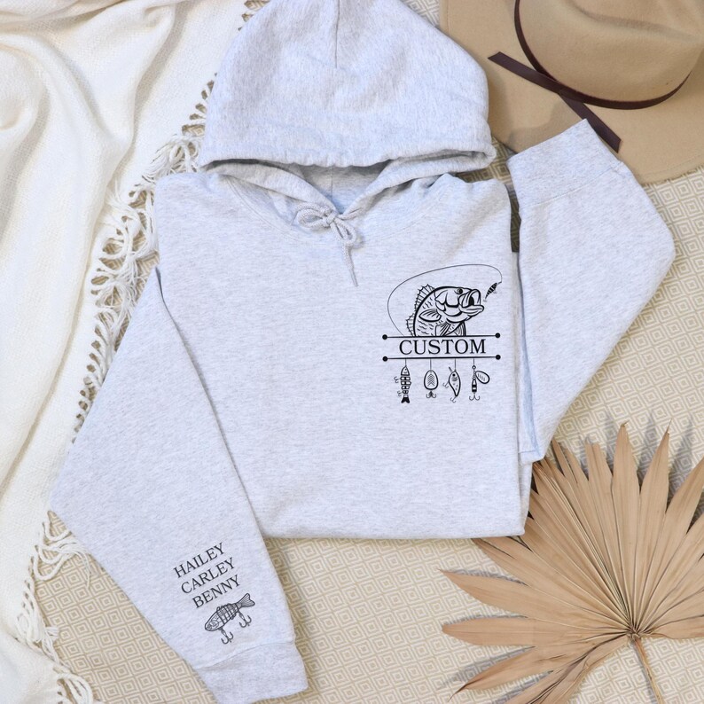 Pode incluir: Camisola de moletom cinza com capuz com um gr&aacute;fico preto de um peixe em uma linha de pesca com a palavra "CUSTOM" e outros iscas de pesca. A manga tem os nomes "HAILEY, CARLEY, BENNY" e um gr&aacute;fico de um peixe.