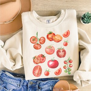 May include: A white sweatshirt with a watercolor design of red tomatoes. The tomatoes are in various sizes and shapes, including whole tomatoes, halved tomatoes, and cherry tomatoes.