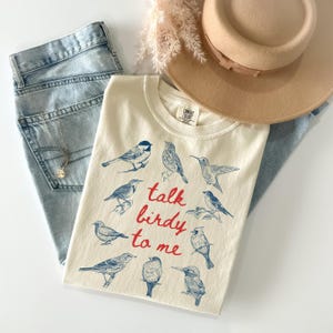 May include: A cream-colored t-shirt featuring a blue ink illustration of nine different birds and the red text "talk birdy to me" printed in the center. The shirt is paired with light blue denim jeans, a tan wide-brimmed hat, and a silver necklace.