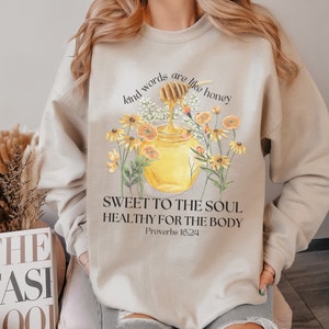 Puede incluir: Sudadera beige con un gr&aacute;fico de un tarro de miel y flores. El texto dice "Kind words are like honey, Sweet to the soul, Healthy for the body, Proverbs 16:24".