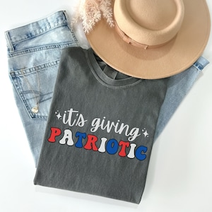 Pu&ograve; includere: Una maglietta grigia con la scritta "it's giving PATRIOTIC" in rosso, bianco e blu. La maglietta &egrave; abbinata a jeans chiari, un cappello beige e una collana d'oro. Gli articoli sono disposti su una superficie bianca.