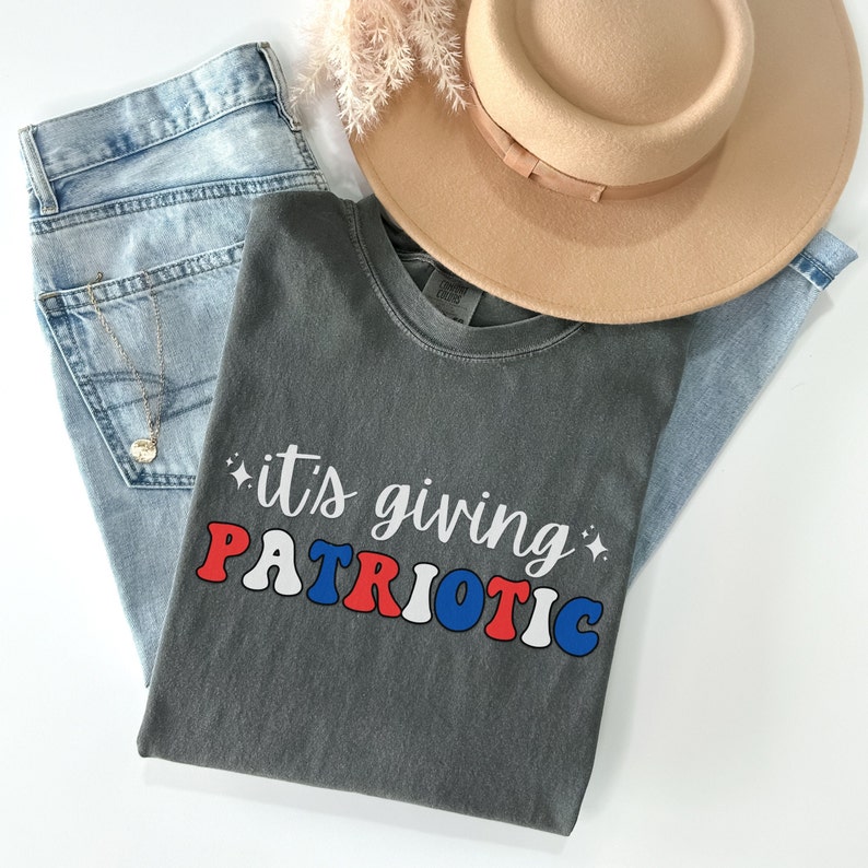 Pu&ograve; includere: Una maglietta grigia con la scritta "it's giving PATRIOTIC" in rosso, bianco e blu. La maglietta &egrave; abbinata a jeans chiari, un cappello beige e una collana d'oro. Gli articoli sono disposti su una superficie bianca.
