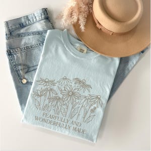 May include: Light blue t-shirt with a graphic of white daisies and the text "Fearfully and Wonderfully Made".
