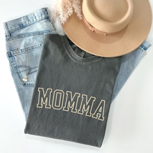 May include: A gray t-shirt with the word "MOMMA" printed in a gold outline on the front. The shirt is folded and laid on top of a pair of blue jeans and a brown hat.