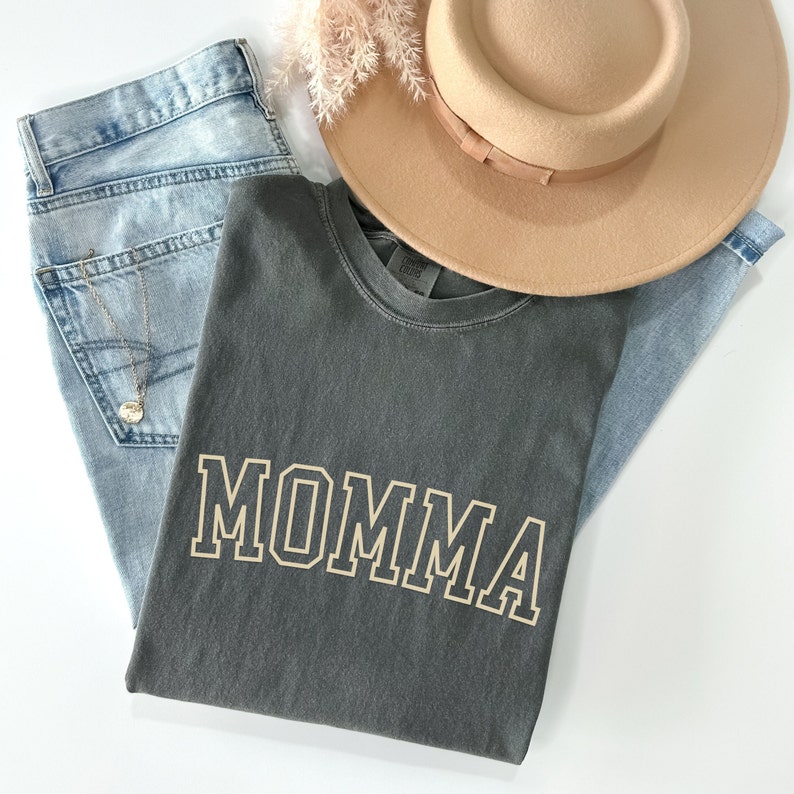 May include: A gray t-shirt with the word "MOMMA" printed in a gold outline on the front. The shirt is folded and laid on top of a pair of blue jeans and a brown hat.
