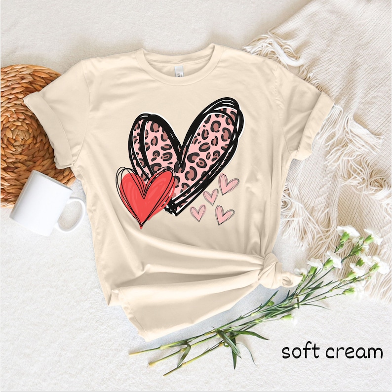 May include: A soft cream colored t-shirt with a graphic of two hearts. The larger heart is pink with a leopard print and the smaller heart is red. There are smaller pink hearts scattered around the larger hearts.