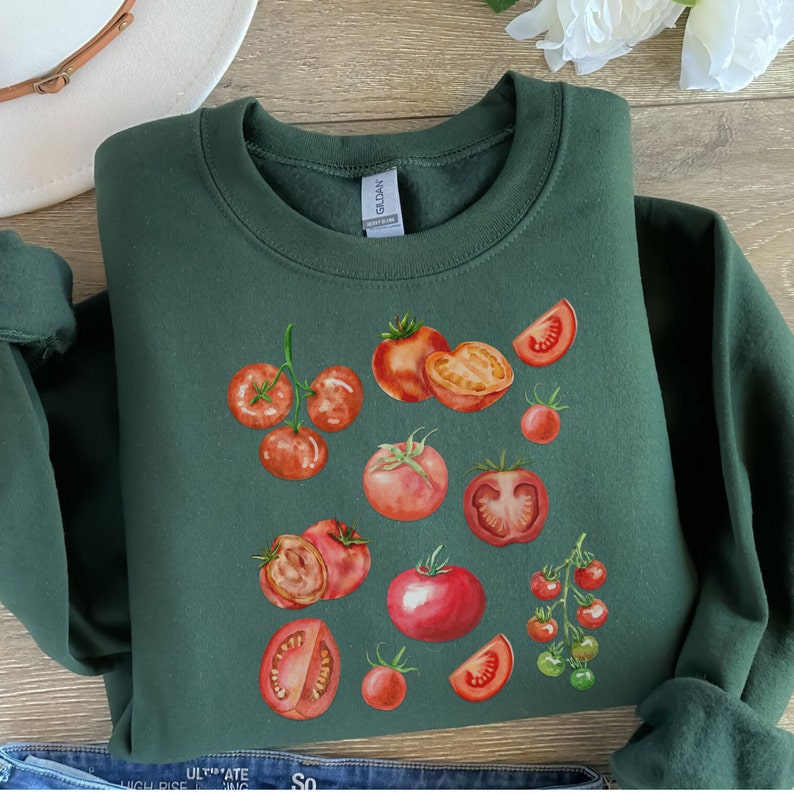 May include: A dark green sweatshirt with a watercolor illustration of red tomatoes. The tomatoes are arranged in a cluster and include whole tomatoes, tomato slices, and cherry tomatoes.