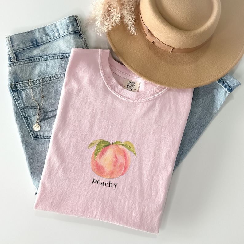 May include: A pink t-shirt with a peach graphic and the word "peachy" printed on the front. The shirt is laid out on a white surface with a pair of blue jeans and a brown hat.