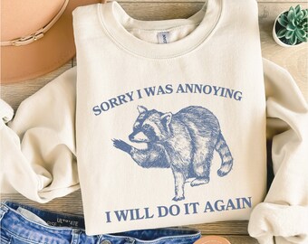 May include: A cream-colored sweatshirt with a blue raccoon graphic and the text "Sorry I was annoying I will do it again".