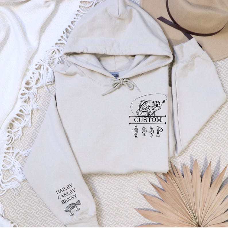 Pode incluir: Um moletom bege com um gr&aacute;fico preto de um peixe em uma linha de pesca com a palavra "CUSTOM" impressa abaixo. O moletom tem um bolso na manga esquerda com os nomes "HAILEY, CARLEY, BENNY" e um pequeno gr&aacute;fico de peixe impresso abaixo.