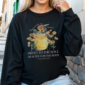 Puede incluir: Sudadera negra con un gr&aacute;fico de un tarro de miel y flores. El texto dice "Kind words are like honey, Sweet to the soul, Healthy for the body, Proverbs 16:24".