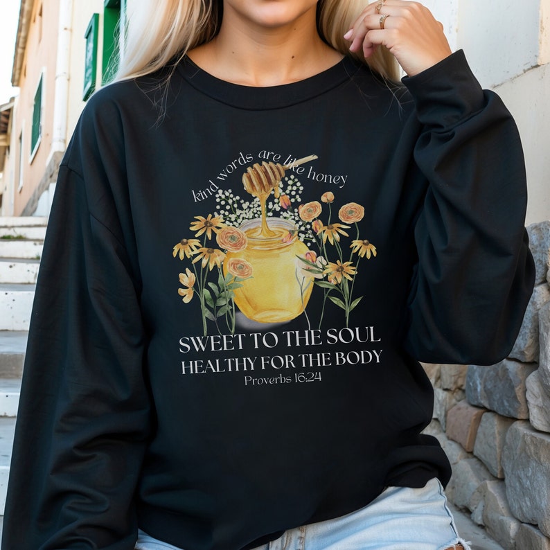 Puede incluir: Sudadera negra con un gr&aacute;fico de un tarro de miel y flores. El texto dice "Kind words are like honey, Sweet to the soul, Healthy for the body, Proverbs 16:24".
