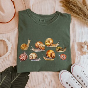 May include: A green t-shirt with a print of multiple snails in various colors and patterns. The snails are arranged in a cluster on the front of the shirt.
