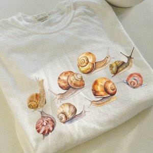 May include: A white t-shirt with a design of eight brown snails with different shell patterns. The snails are arranged in a cluster on the shirt.