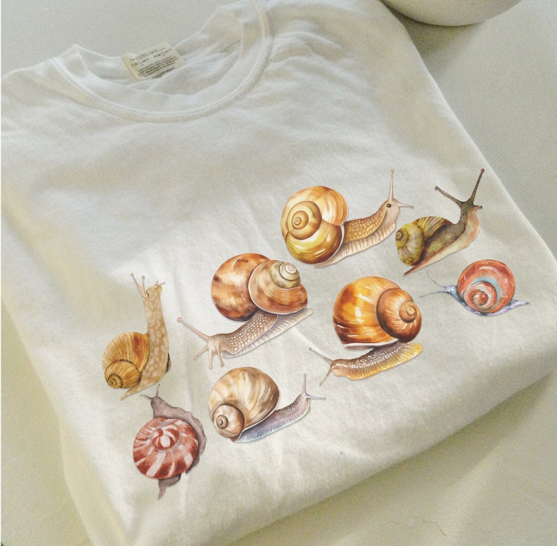 May include: A white t-shirt with a design of eight brown snails with different shell patterns. The snails are arranged in a cluster on the shirt.