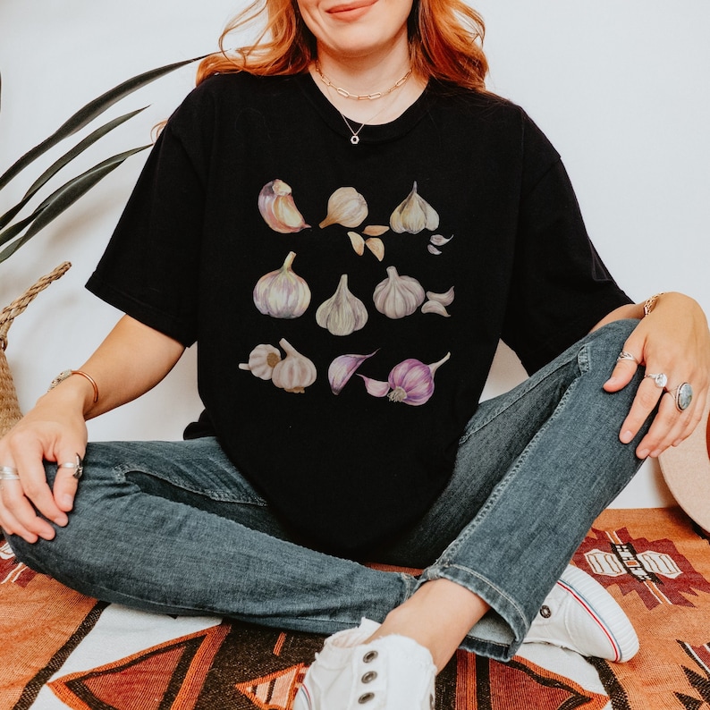 May include: A black t-shirt with a graphic print of various garlic cloves in shades of purple, white, and brown. The shirt is being worn by a person with long brown hair.