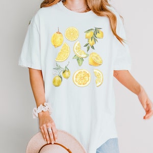 Pu&ograve; includere: Una maglietta bianca con una stampa di limone. I limoni sono gialli e verdi e sono disposti in un motivo sparso sulla maglietta.