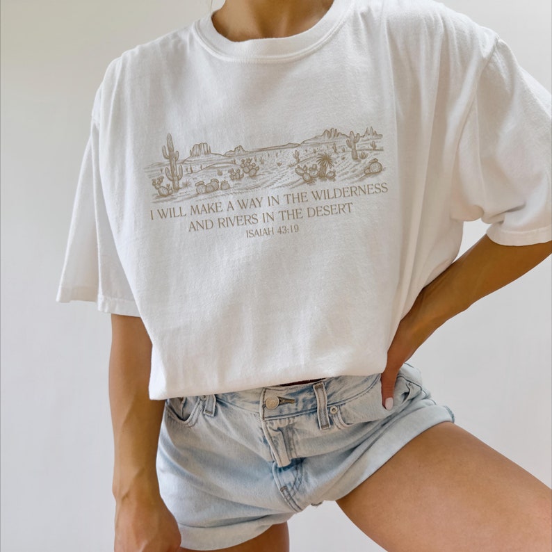 Puede incluir: Camiseta blanca con un dise&ntilde;o de paisaje des&eacute;rtico en tonos marrones. El dise&ntilde;o incluye cactus, monta&ntilde;as y el texto "I WILL MAKE A WAY IN THE WILDERNESS AND RIVERS IN THE DESERT ISAIAH 43:19". La camiseta tiene mangas cortas y cuello redondo.