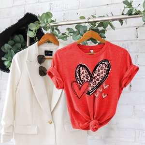 May include: Red t-shirt with a graphic of two hearts. The larger heart is pink and has a leopard print. The smaller heart is red. There are smaller red hearts around the larger hearts.