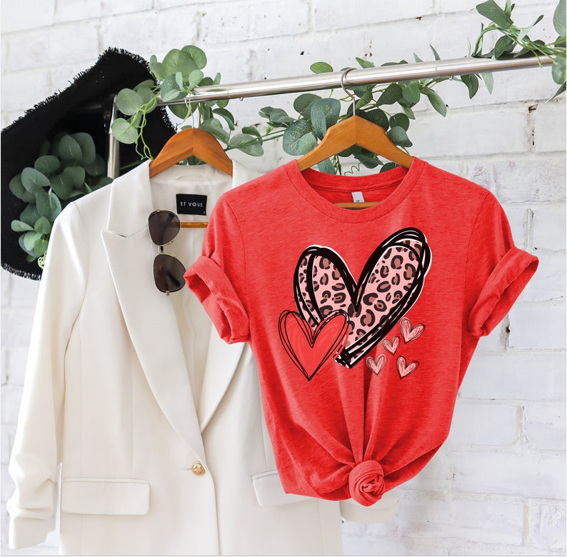 May include: Red t-shirt with a graphic of two hearts. The larger heart is pink and has a leopard print. The smaller heart is red. There are smaller red hearts around the larger hearts.