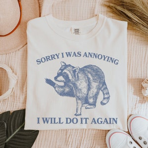 May include: White t-shirt with a blue illustration of a raccoon and the text "Sorry I was annoying I will do it again".