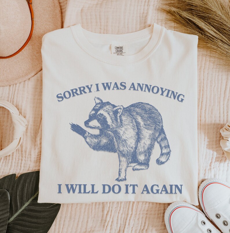 May include: White t-shirt with a blue illustration of a raccoon and the text "Sorry I was annoying I will do it again".