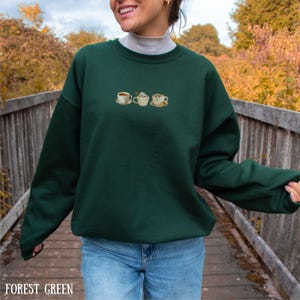 May include: Forest green sweatshirt with embroidered coffee cup designs. The sweatshirt features a crew neck and long sleeves. The text "FOREST GREEN" is displayed at the bottom of the image.