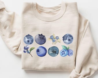 May include: A beige sweatshirt with a watercolor design of blueberries and leaves. The blueberries are various shades of blue and purple.