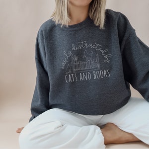 May include: A dark gray sweatshirt with a white graphic design that says "easily distracted by cats and books". The graphic features a bookshelf with cats sitting on top of the books.